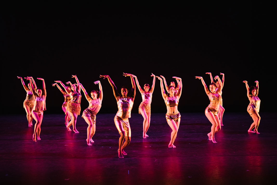 A row of college student dancers in shiny gold outfits raise their hands on stage